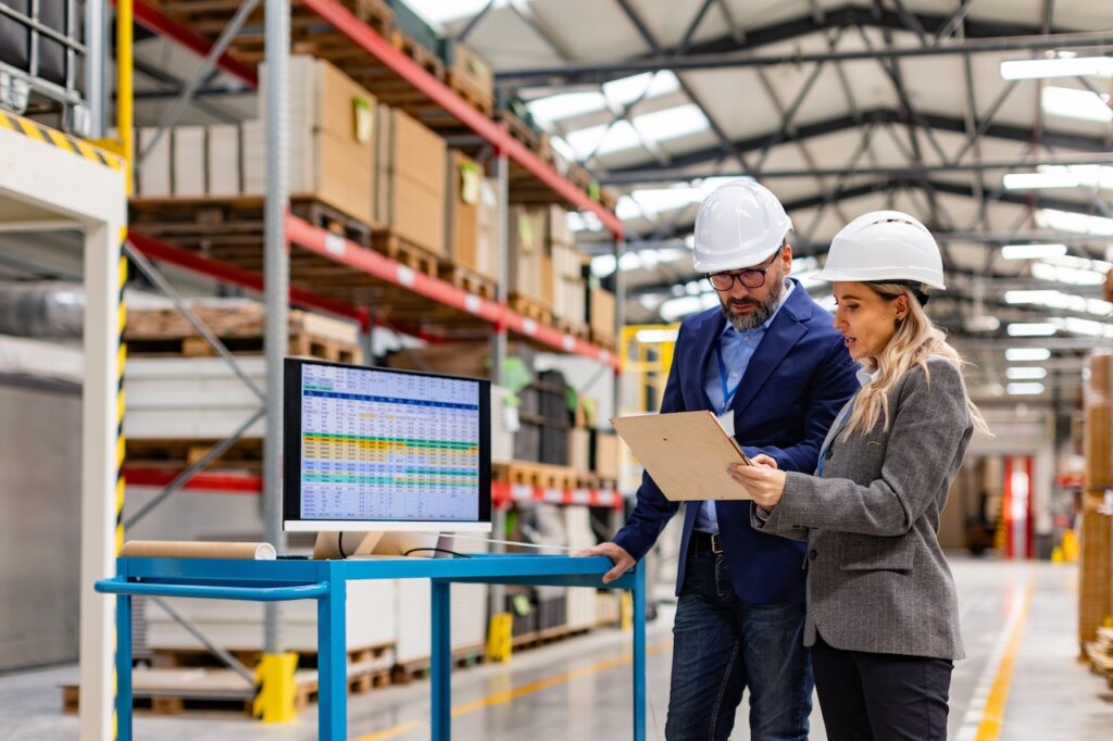 Zwei Ingenieure mit Schutzhelmen überprüfen Produktionsdaten auf einem Clipboard, während ein Computer mit Bestandsmanagementsoftware in einem großen Lager steht.